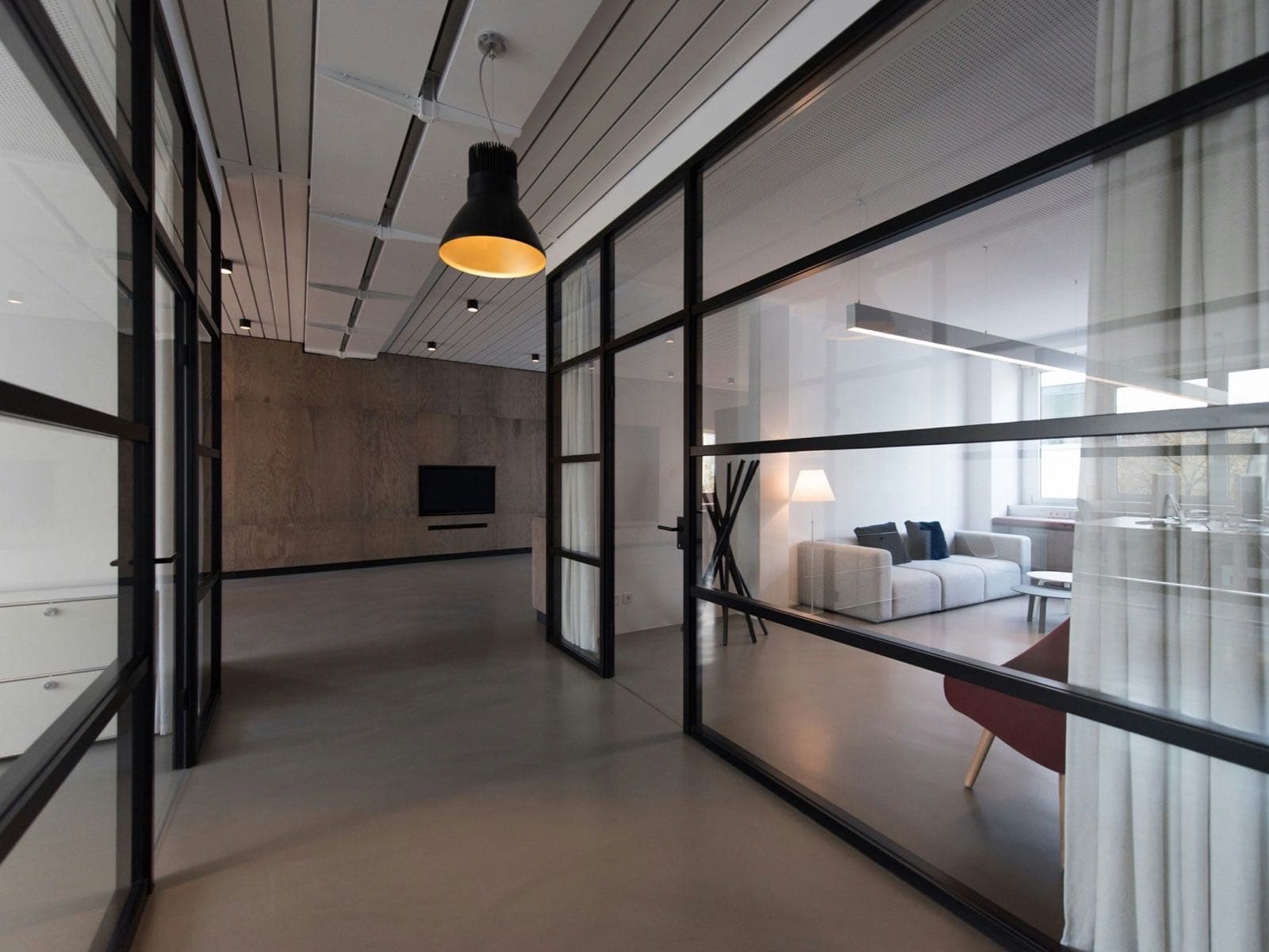Un couloir vitré de bureaux : cloisons d'acier noir, suspension allumée, un salon d'attente derrière la vitre.